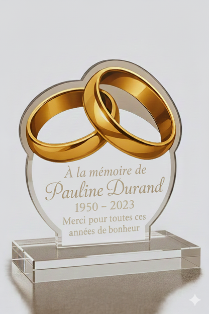 Plaque funéraire personnalisée en plexiglas transparent avec deux alliances dorées entrelacées, gravure élégante avec prénom, dates et message de souvenir, finition haut de gamme et design moderne.